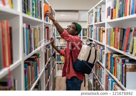 Nerd student african american man choosing book in university library taking it from shelf. Nerd student african american man choosing book in university library taking it from shelf. 121678892