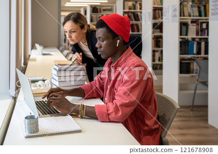 Interested african american student typing on laptop near focused college teacher looking at screen Interested african american student typing on laptop near focused college teacher looking at screen 121678908