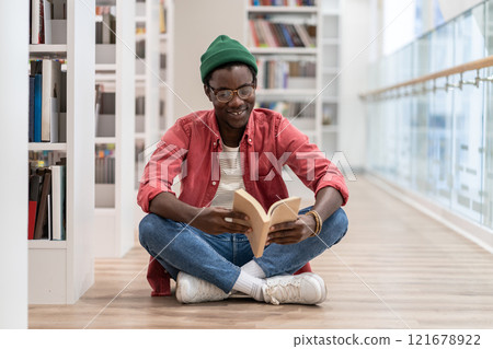 Happy African American millennial guy sitting on floor reading book while visiting library 121678922