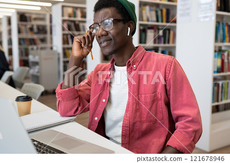 Positive inspired African American guy student looking at laptop screen sits in library or bookstore Positive inspired African American guy student looking at laptop screen sits in library or bookstore 121678946