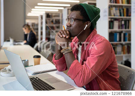 Thoughtful black male student wearing wireless earbuds sitting at desk with laptop in library 121678966