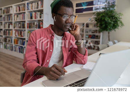 Thoughtful hipster man student or freelancer thinks of online project looking at laptop in library. Thoughtful hipster man student or freelancer thinks of online project looking at laptop in library. 121678972