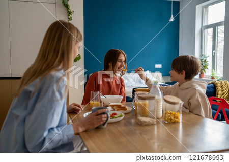 Smiling kid brother shares cornflakes with older teen sister during fun family breakfast with mother Smiling kid brother shares cornflakes with older teen sister during fun family breakfast with mother 121678993