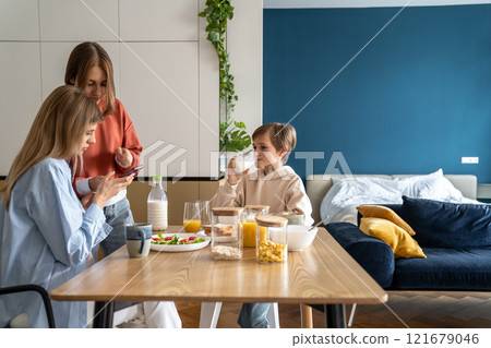 Interested teen girl show to mom news on cellphone while brother drink glass milk breakfast at table Interested teen girl show to mom news on cellphone while brother drink glass milk breakfast at table 121679046