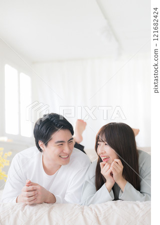 Smiling couple looking at each other while lying face down on a bed in a bedroom Smiling couple looking at each other while lying face down on a bed in a bedroom 121682424