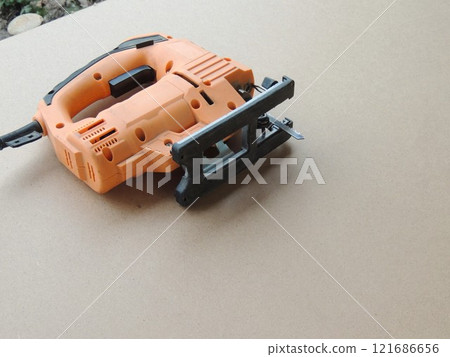 orange corded electric jigsaw lying on light brown sheet of pressed wood or plywood as abstract background with working tool of carpenter or decorator craftsmanship without people 121686656
