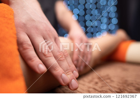 Unrecognizable satisfied woman lies on table, male masseur touches and kneads legs of patient covered with soft towel and resting 121692367