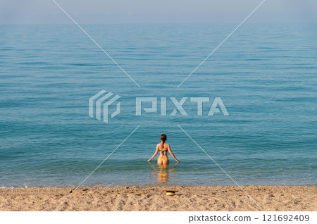 A young Caucasian woman in a different swimsuit and stands waist-deep in sea water on a sunny day. Her back is to the camera 121692409