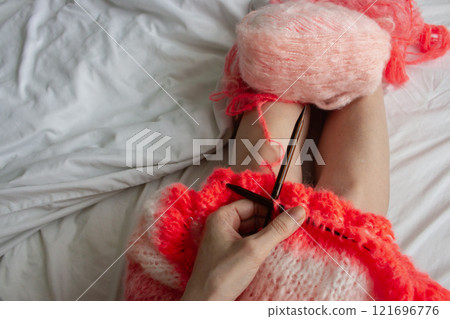Woman with coral yarn and wooden knitting needles on white bed sheet, top view. Women legs and bright knitted sweater, view from above. Knitting hobby. Cozy home activity. Creative lifestyle. Woman with coral yarn and wooden knitting needles on white bed sheet, top view. Women legs and bright knitted sweater, view from above. Knitting hobby. Cozy home activity. Creative lifestyle. 121696776
