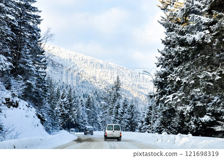 A winter road passing through a snow-covered spruce forest, with several cars driving along it. 121698319
