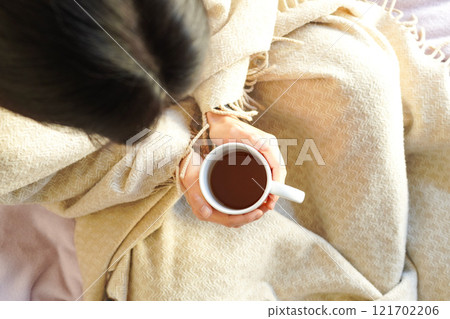A young woman wrapped in a blanket in bed at home and holding a mug of hot drink in both hands, overhead view 121702206