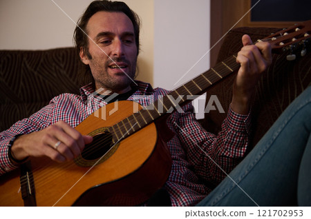 Man Playing Acoustic Guitar While Relaxing in a Comfortable Indoor Setting 121702953