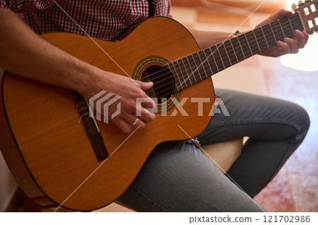 Person playing a classic guitar while seated on a casual indoor setting 121702986