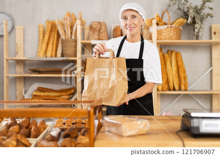 Senior woman entrepreneur owner of bakery holds out paper bag with pastries in hand Senior woman entrepreneur owner of bakery holds out paper bag with pastries in hand 121706907
