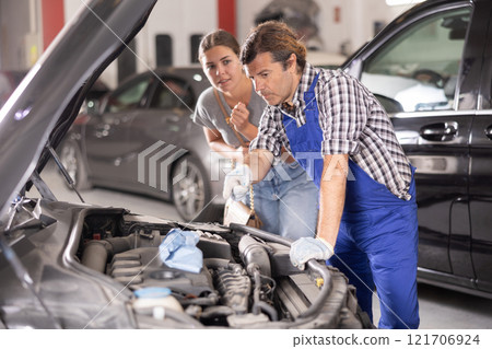 Woman talking to auto mechanic in uniform about repairing his car engine in auto repair shop Woman talking to auto mechanic in uniform about repairing his car engine in auto repair shop 121706924
