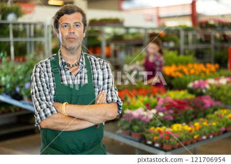 Man worker stands in aisle between rows with variety of flowering plants in pots 121706954
