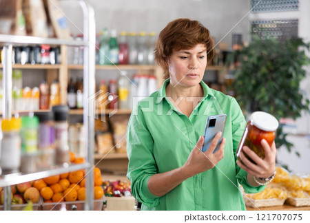 Checking expiration date of canned food - female shopper scans QR code on label of pickled tomatoes using smartphone 121707075