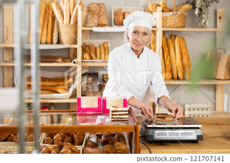 Elderly female seller weighs piece of sweet cake on scales in the interior of private bakery Elderly female seller weighs piece of sweet cake on scales in the interior of private bakery 121707141