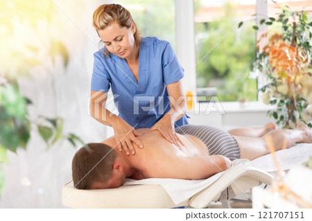 Female masseur performs back stroking and patting during massage session for male client 121707151