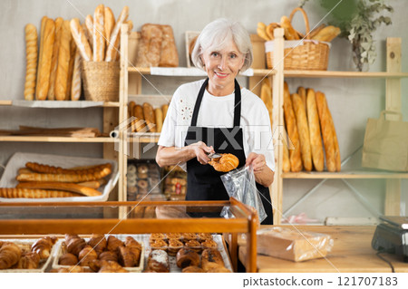 Senior woman seller puts croissant in plastic disposable bag for buyer, packs goods. Senior woman seller puts croissant in plastic disposable bag for buyer, packs goods. 121707183