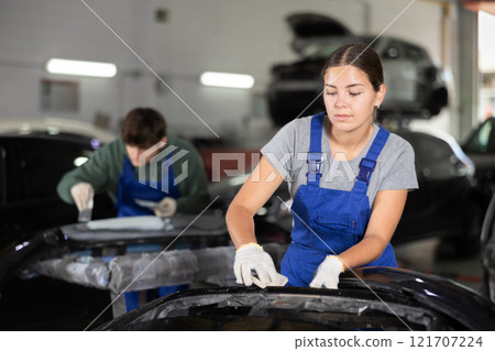 Woman mechanic prepares car for painting in workshop - grinds and puts putty on bumper before painting 121707224