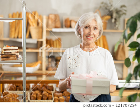 Positive elderly woman bought cake in bakery, packed in white elegant box with pink bow Positive elderly woman bought cake in bakery, packed in white elegant box with pink bow 121707254