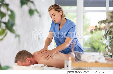 Female masseur performs back stroking and patting during massage session for male client Female masseur performs back stroking and patting during massage session for male client 121707257