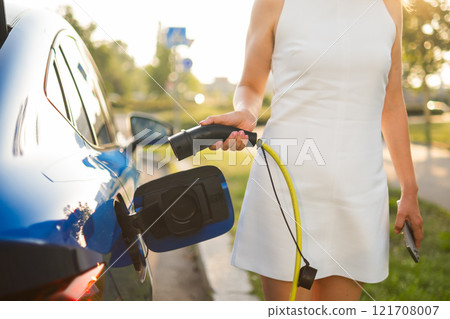 Woman connects a charging cable to her electric car in a bright, green area during the day. Woman connects a charging cable to her electric car in a bright, green area during the day. 121708007