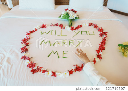 Woman hand wearing a diamond ring after say YES with message WILL YOU MARRY ME made of flower petals with bouquet on bed. Idea for marriage proposal, romantic surprise, wedding and engagement concept 121710507