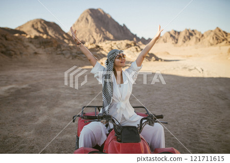 A rather attractive woman is going on a safari, driving an ATV in the desert. Beautiful young woman with a scarf on her head sits on a red four-wheeled ATV and rides towards adventure 121711615