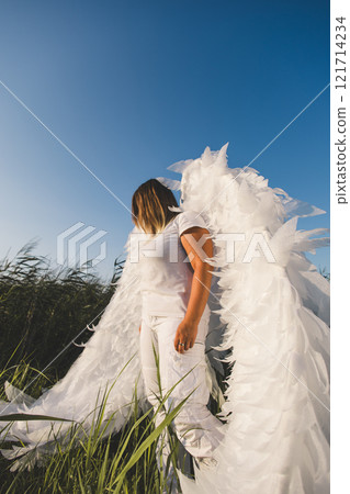 An ethereal angel stands against a blue sky. A woman with large white angel wings stands in a field of tall grass, against a clear blue sky, creating a serene and ethereal atmosphere. 121714234