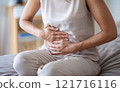Woman in White Blouse Experiencing Abdominal Pain While Sitting on Sofa at Home 121716116