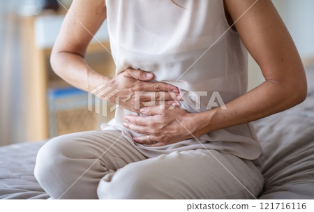 Woman in White Blouse Experiencing Abdominal Pain While Sitting on Sofa at Home Woman in White Blouse Experiencing Abdominal Pain While Sitting on Sofa at Home 121716116