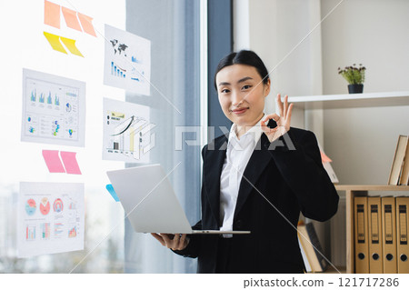 Confident Asian businesswoman holding laptop, presenting data charts at modern office setting with positive gesture, boosting success and professionalism. Confident Asian businesswoman holding laptop, presenting data charts at modern office setting with positive gesture, boosting success and professionalism. 121717286