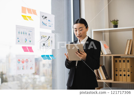 Asian businesswoman in formal attire analyzing data charts on digital tablet in modern office. Reflective charts on window indicate strategic planning and productivity. 121717306