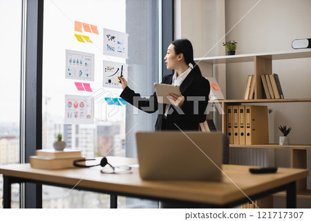 Young Asian businesswoman in office analyzes data charts on glass wall. She uses tablet for insights, exemplifying modern work. Professional setting with city view enhances focus. 121717307