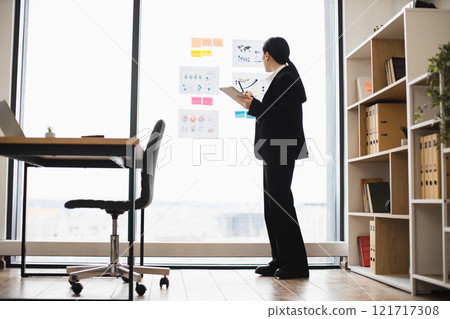 Asian businesswoman studying charts on window with clipboard. Professional setting in modern office environment with daylight. 121717308