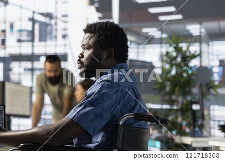 Person with paraplegia sitting in wheelchair at desk in office, doing research for team project. Man with disability looking at computer screen in inclusive workplace, solving tasks Person with paraplegia sitting in wheelchair at desk in office, doing research for team project. Man with disability looking at computer screen in inclusive workplace, solving tasks 121718508