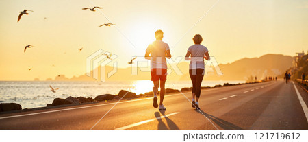 Couple jogging along a scenic coastal road at sunset near a tranquil beach with seagulls flying above 121719612