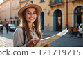 A smiling young woman in a hat with a backpack holds a map in a sunlit European town square. 121719715