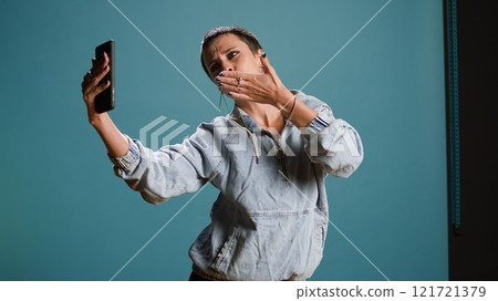 Silly confident woman recording a video while she shows groovy dance moves, filming on her smartphone and fooling around in studio. Funky bold edgy person being funny. Camera A. 121721379