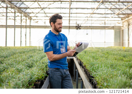 Young bearded man in a blue thisrt standing in a greenhouse and looking enjoyed 121721414