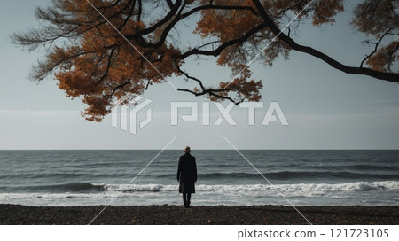 A man stands on the seashore under a large autumn tree and looks at the horizon 121723105