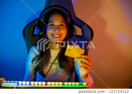 Asian gamer playing online video game and holding a credit card for payment game online, young woman in gaming headphones using computer for playing games at home, broadcast streaming live, Front view 121724312