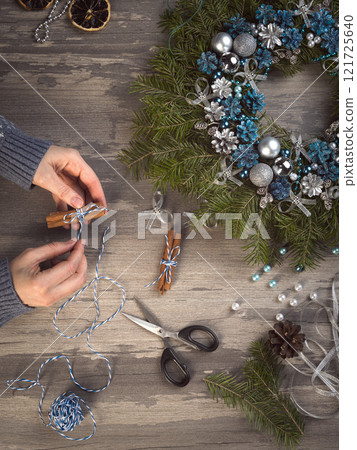 Process of making Christmas decoration. Female hands with cinnamon sticks next to blue Christmas wreath, scissors, beads, cones, twine on a wooden background, vertical image. 121725640