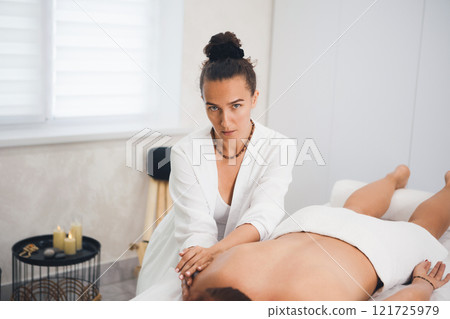 Attractive masseuse in white outfit gently massages a lying woman in pro salon, copy space, selective focus. 121725979