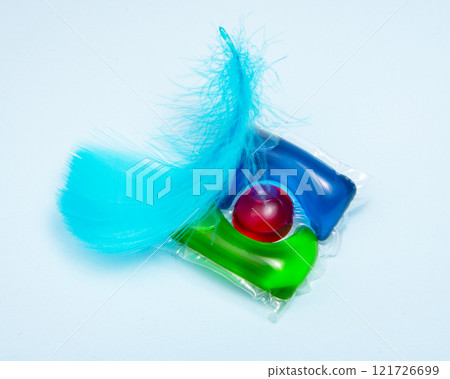 Liquid powder in gel capsule for washing multicolor with blue feather on color background Liquid powder in gel capsule for washing multicolor with blue feather on color background 121726699