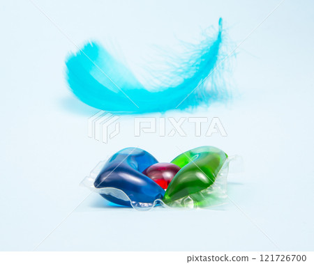 Liquid powder in gel capsule for washing multicolor with blue feather on color background Liquid powder in gel capsule for washing multicolor with blue feather on color background 121726700