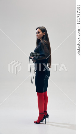 Side view image of beautiful businesswoman in black blouse, skirt and red tights with heeled shoes, posing with small bag in hands against studio background Side view image of beautiful businesswoman in black blouse, skirt and red tights with heeled shoes, posing with small bag in hands against studio background 121727195