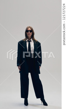 Elegant and stylish businesswoman wear total black suit with white shirt, tie and sunglasses, posing with confidence against studio background Elegant and stylish businesswoman wear total black suit with white shirt, tie and sunglasses, posing with confidence against studio background 121727224